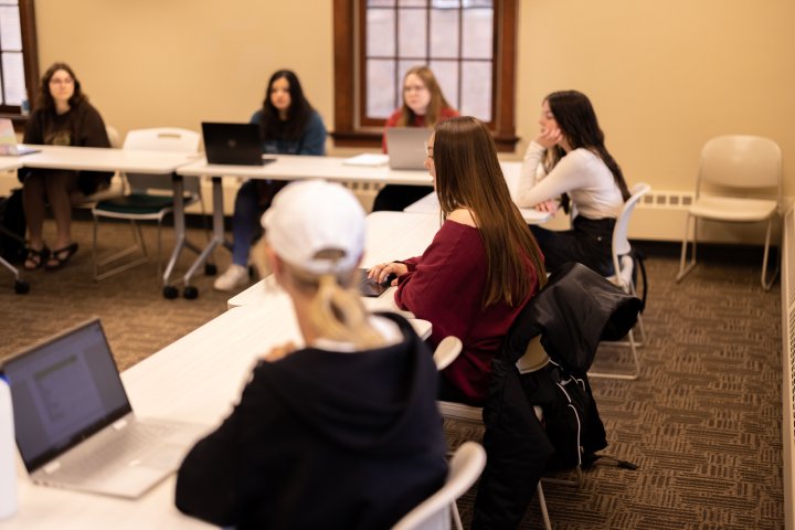 A group of students sits around tables arranged in a U-shape, engaging in a classroom discussion. Some students have laptops open in front of them, and a window is visible in the background. The atmosphere appears casual and focused.
